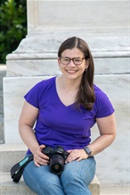 white young woman, dark brown hair, pulled back into a lower ponytail. She's wearing a brown long sleeved shirt and a book bag strap across her body. She's posed in front of the White House at night smiling.