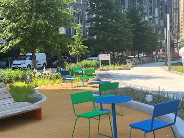 A sunlit urban park plaza featuring vibrant green and blue metal patio chairs and round tables arranged along a winding tan walkway.
