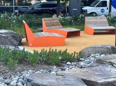 A sunlit urban park plaza featuring vibrant patio chairs arranged along a winding tan walkway.