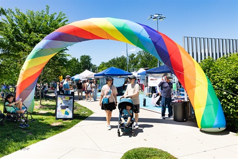 A large, inflatable rainbow arch frames the entrance to a bright, outdoor community festival on a sunny day. Underneath the arch, a diverse group of people walk along a paved path, including a couple pushing a young child in a stroller. In the background, several blue and white event tents are lined up on a blue-surfaced court, with lush green trees and a clear blue sky visible.