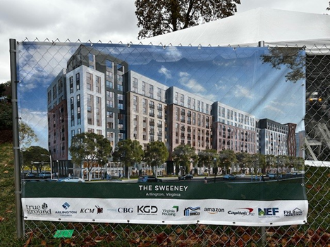 A banner hung on a chain link fence shows a multistory building on a tree-lined street.